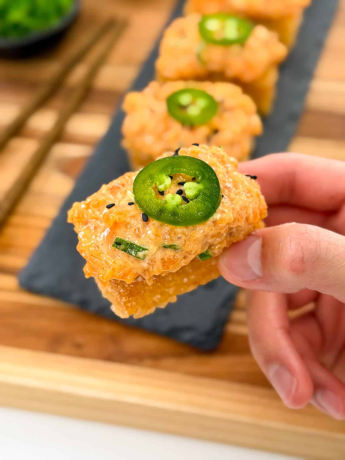 A hand holds a piece of crispy rice topped with spicy tuna, a jalapeño slice, and black sesame seeds. Similar Spicy Salmon Crispy Rice bites are lined up on a slate serving tray in the background. Chopsticks and a bowl are also visible.
