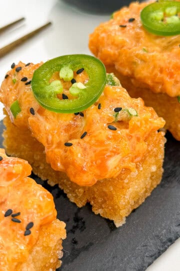 Three pieces of Spicy Salmon Crispy Rice, topped with jalapeño slices and black sesame seeds, are arranged on a slate serving tray, with chopsticks placed nearby.