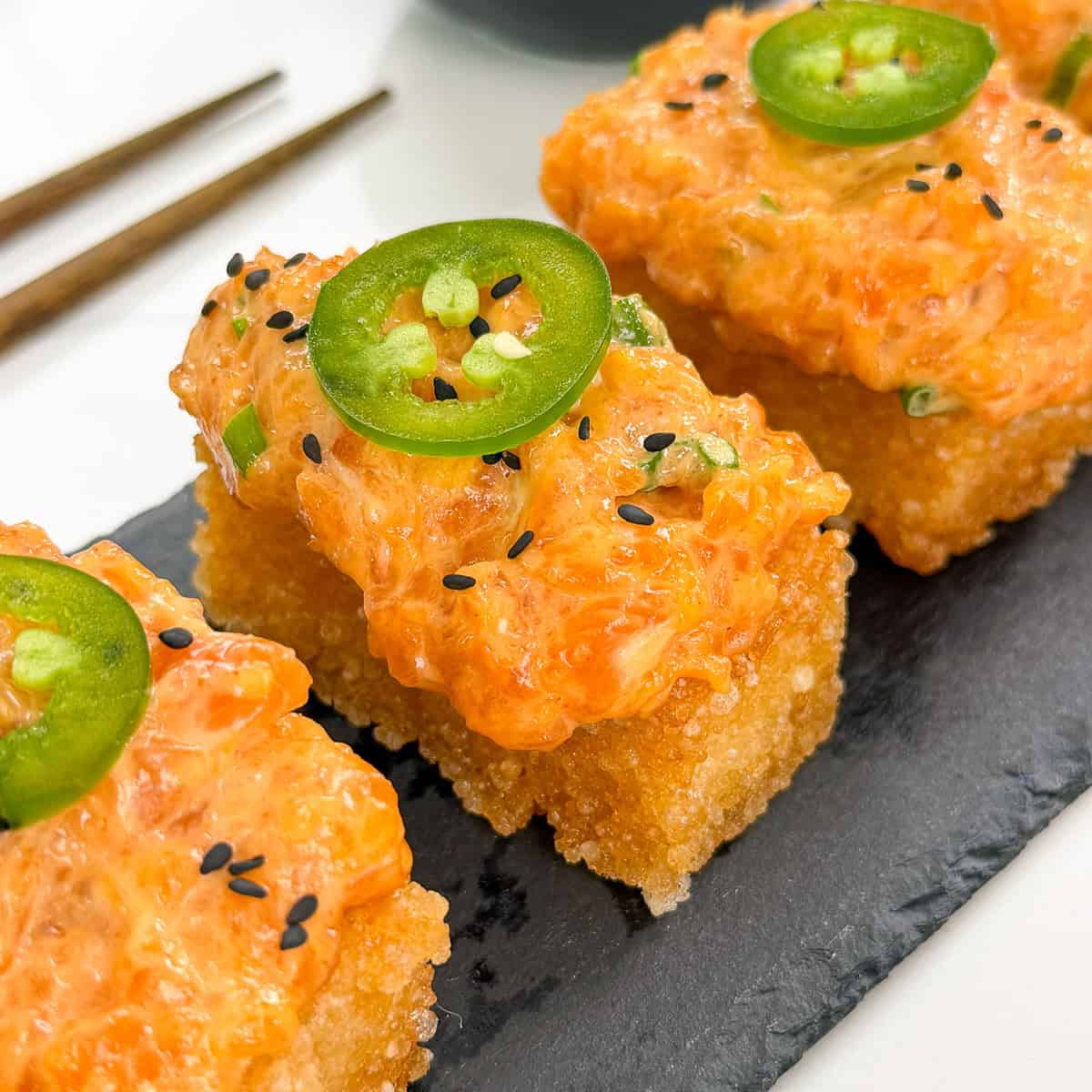 Three pieces of Spicy Salmon Crispy Rice, topped with jalapeño slices and black sesame seeds, are arranged on a slate serving tray, with chopsticks placed nearby.