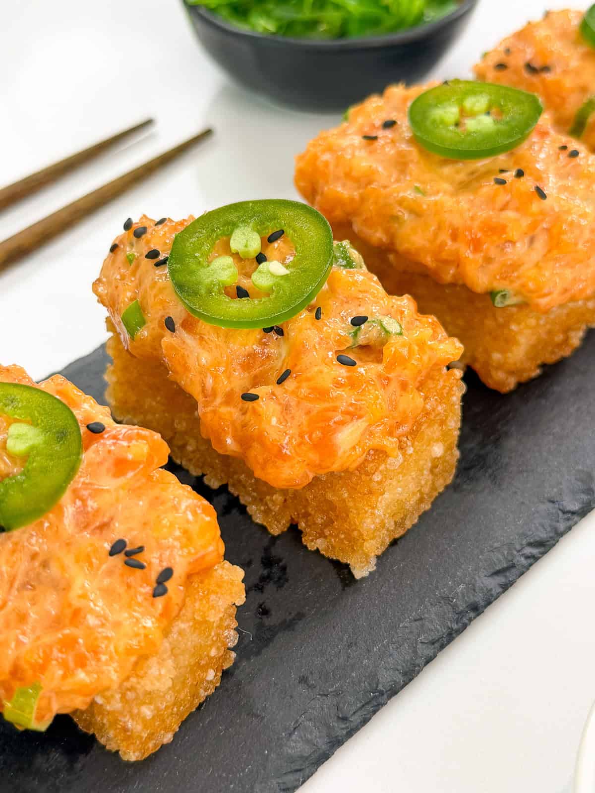 Three pieces of Spicy Salmon Crispy Rice, topped with black sesame seeds and sliced jalapeño, are arranged on a black slate serving board. Chopsticks and a bowl of green seaweed salad sit in the background.