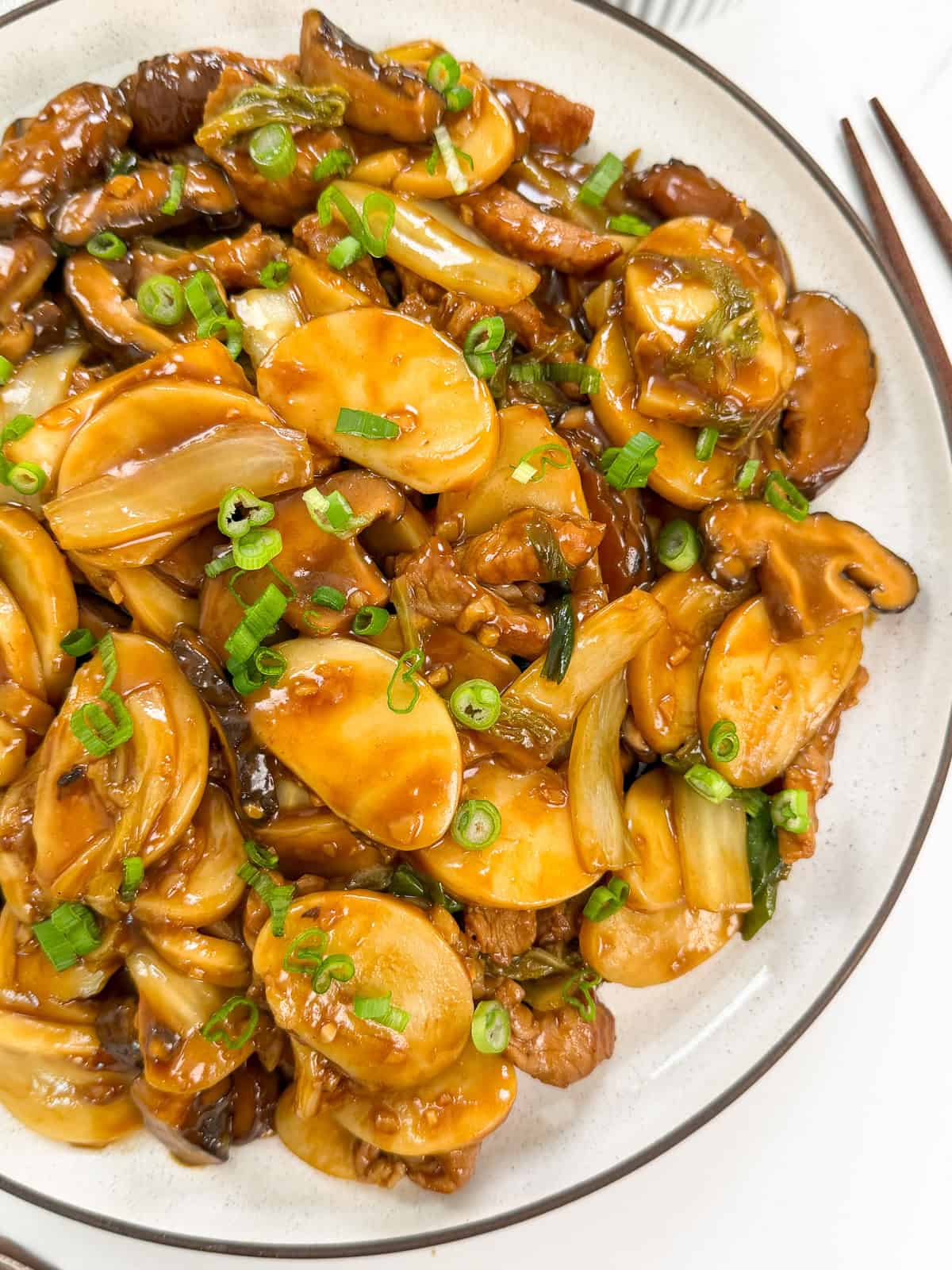 A plate of Stir-Fried Shanghai Rice Cakes (Chao Nian Gao) with sliced mushrooms, green onions, and vegetables tossed in a glossy brown sauce, served on a white dish with chopsticks beside it.