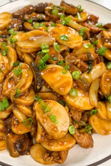 A plate of Stir-Fried Shanghai Rice Cakes (Chao Nian Gao) with sliced mushrooms, beef, leafy greens, and chopped green onions in a glossy brown sauce, served on a white dish.