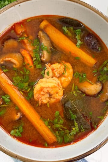 A bowl of Tom Yum Soup Recipe with shrimp, lemongrass stalk, kaffir lime leaves, chopped cilantro on a blue towel
