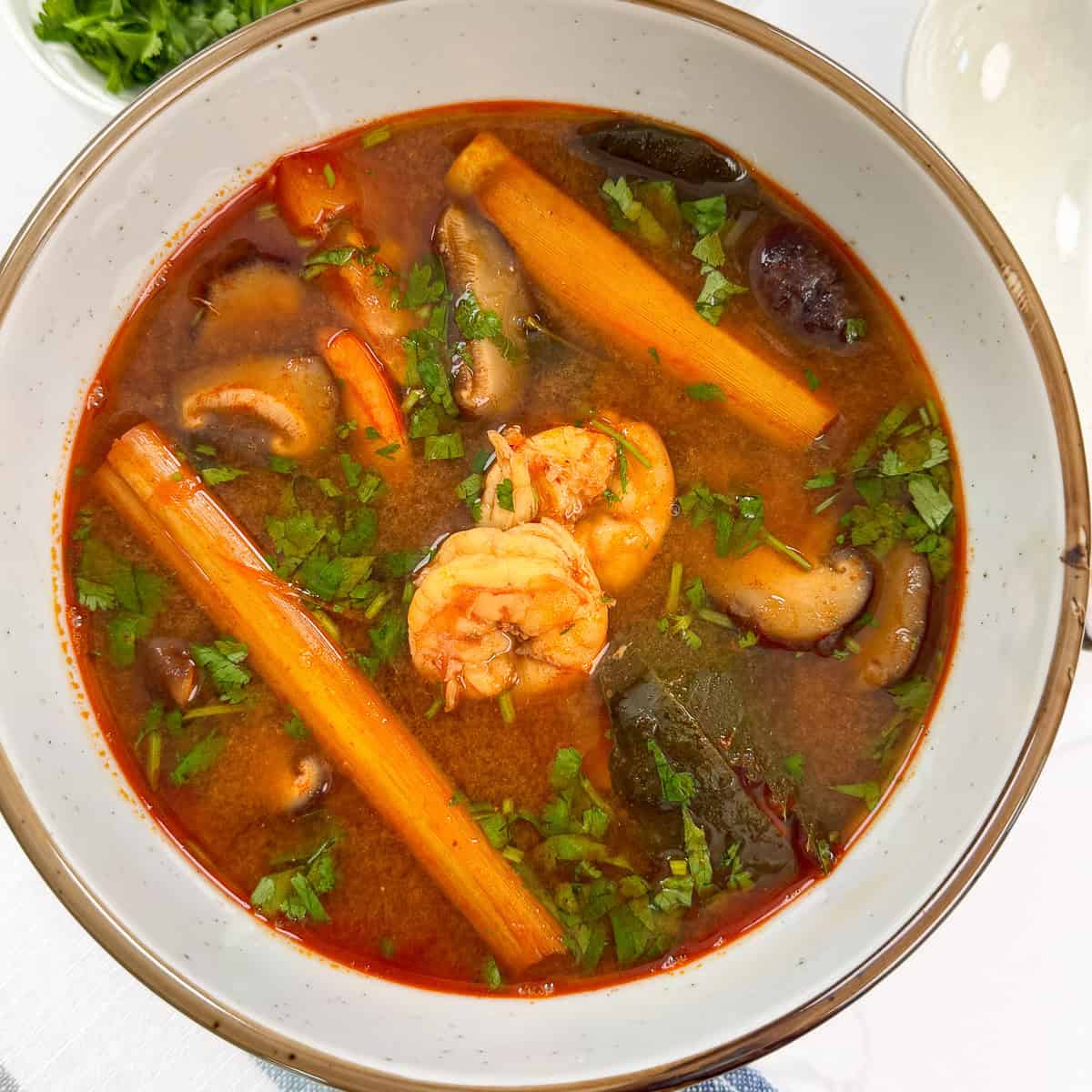 A bowl of Tom Yum Soup Recipe with shrimp, lemongrass stalk, kaffir lime leaves, chopped cilantro on a blue towel
