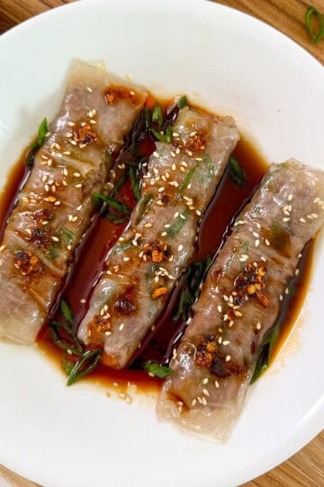 Beef Rice Paper Noodle Rolls (Cheung Fun) on a white plate with soy sauce and sesame seeds