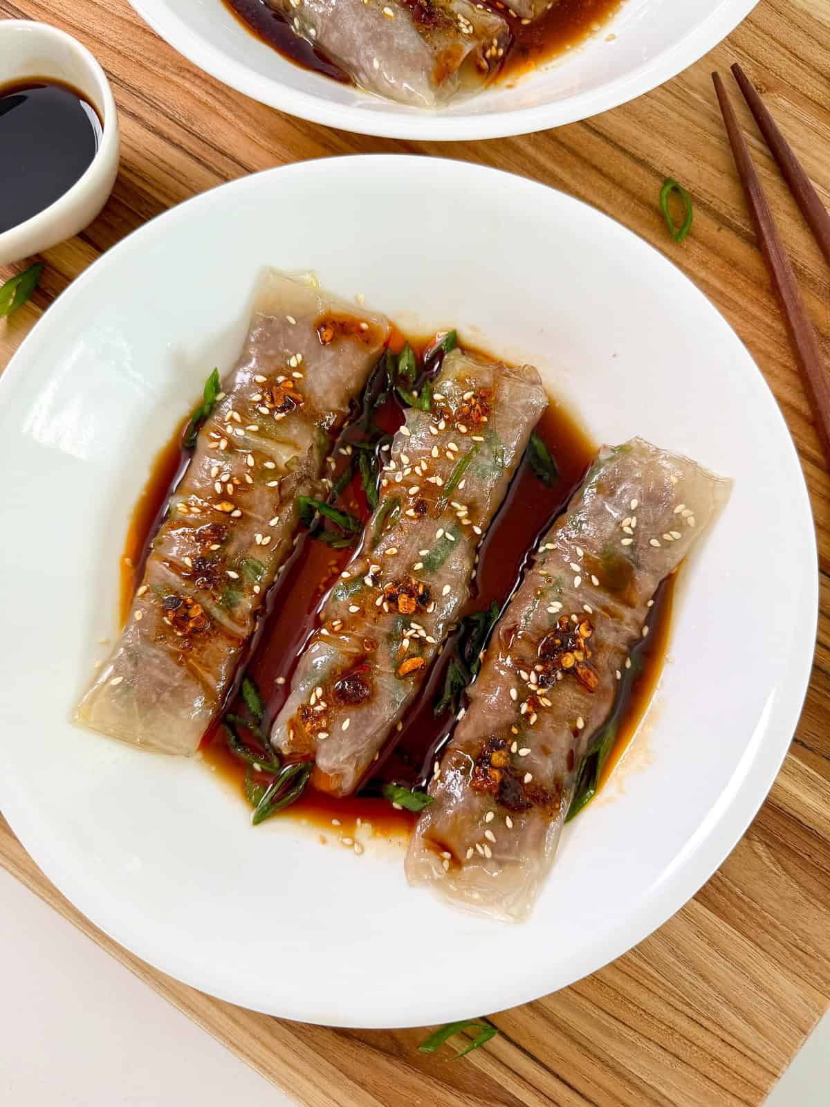 Beef Rice Paper Noodle Rolls (Cheung Fun) on a white plate with soy sauce and sesame seeds. Chopsticks are on the right side of the plate.