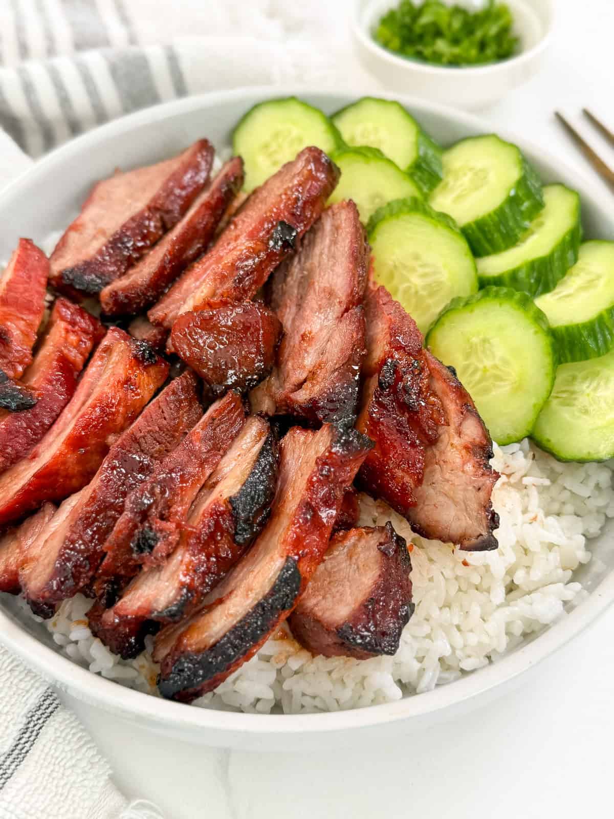 Char siu pork (Chinese BBQ pork) sitting on top of a bed of white rice with cucumbers on the side