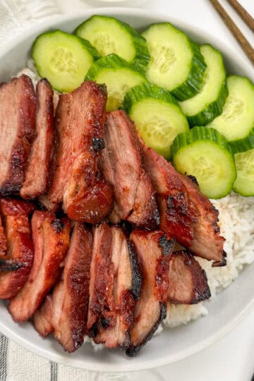 Char Siu Pork (Chinese BBQ Pork) on top of a bed of white rice and side of sliced cucumbers. A fork is on the right and a towel on the left side.