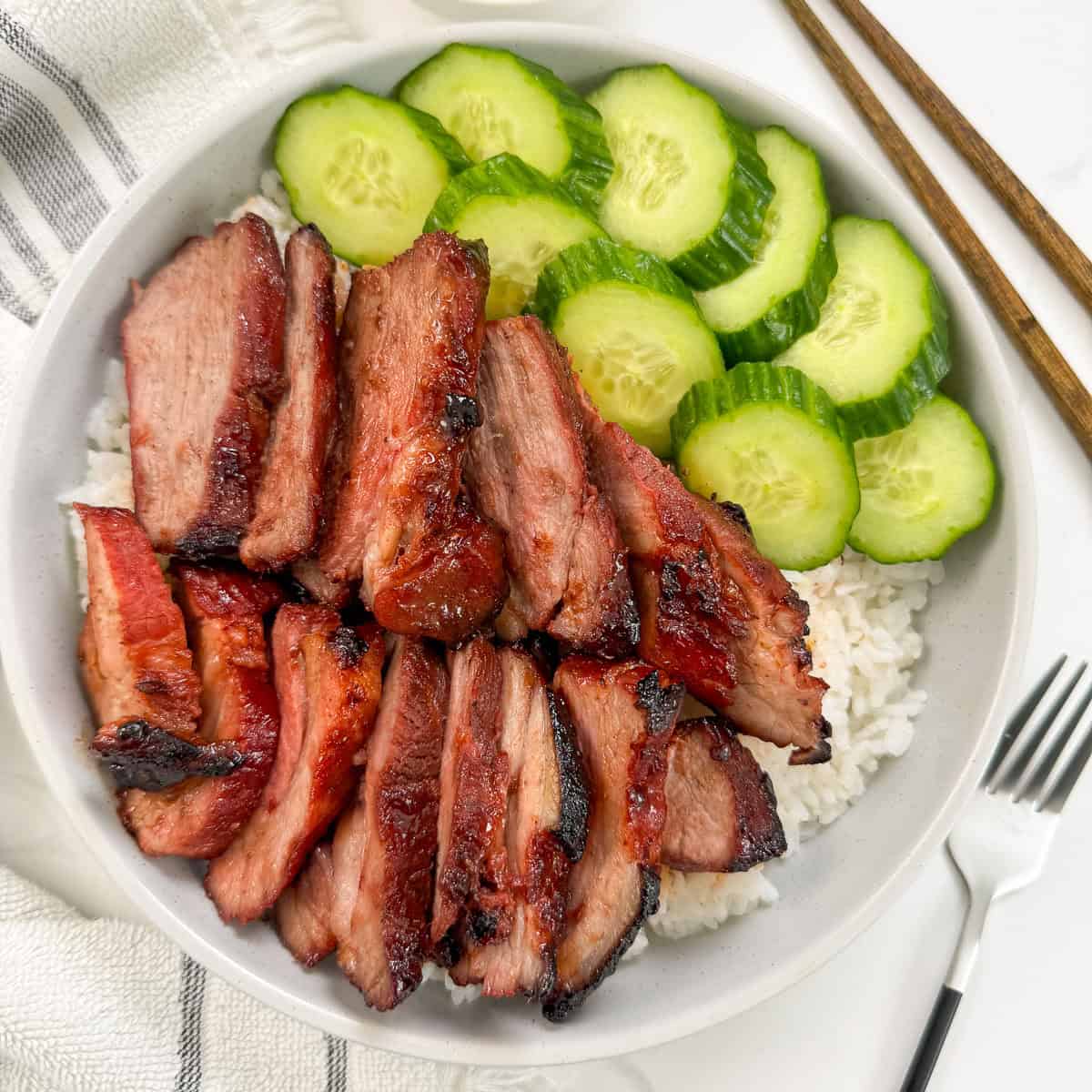 Char Siu Pork (Chinese BBQ Pork) on top of a bed of white rice and side of sliced cucumbers. A fork is on the right and a towel on the left side.