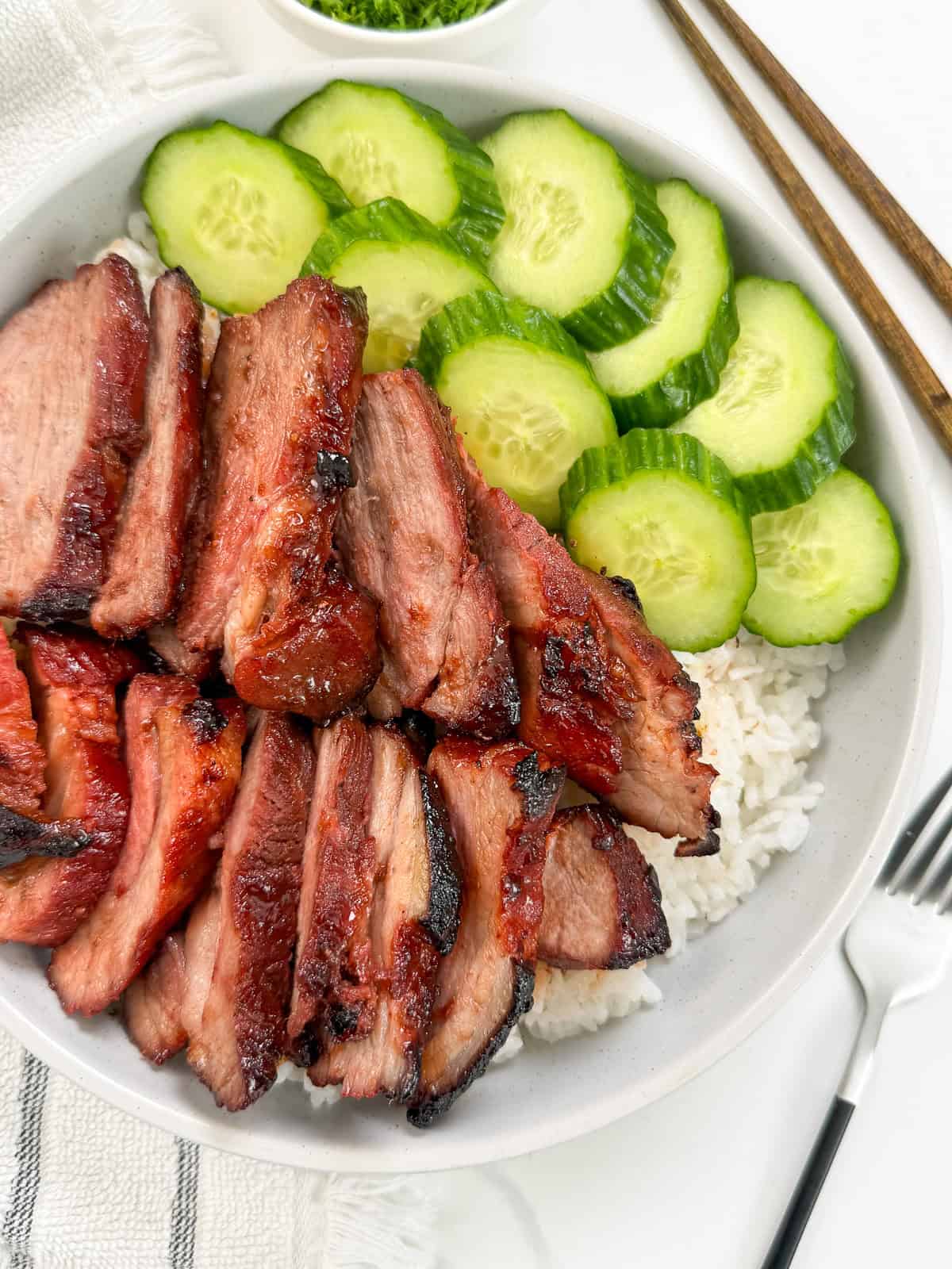 Char Siu Pork (Chinese BBQ Pork) on top of a bed of white rice and side of sliced cucumbers. A fork is on the right, a towel on the left side, and a pair of chopsticks on the top.