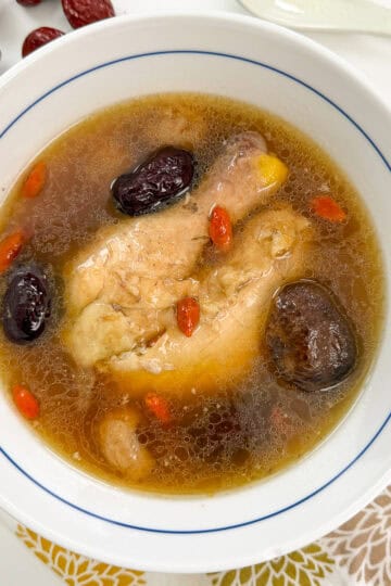 Chinese Chicken Herbal Soup in a white bowl with chicken thighs, shiitake mushrooms, red dates, goji berries. A spoon sits at the top.
