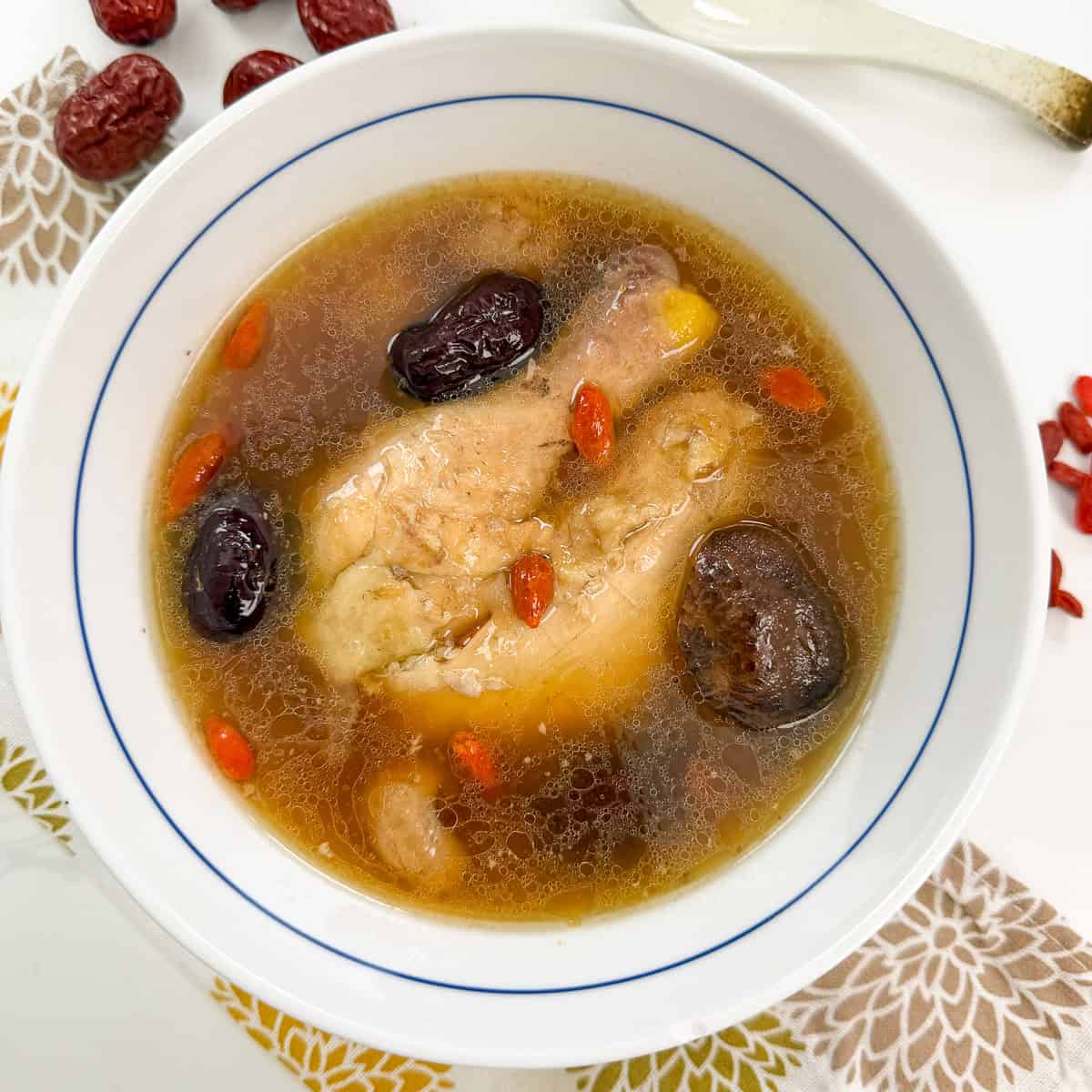 Chinese Chicken Herbal Soup in a white bowl with chicken thighs, shiitake mushrooms, red dates, goji berries. A spoon sits at the top.