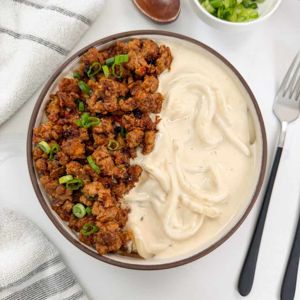 Creamy Mapo Tofu Udon in a bowl with minced meat and green onions on top. There is a fork on the right side and a wooden spoon above the bowl.