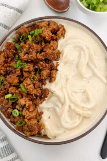 Creamy Mapo Tofu Udon in a bowl with minced meat and green onions on top. There is a fork on the right side and a wooden spoon above the bowl.