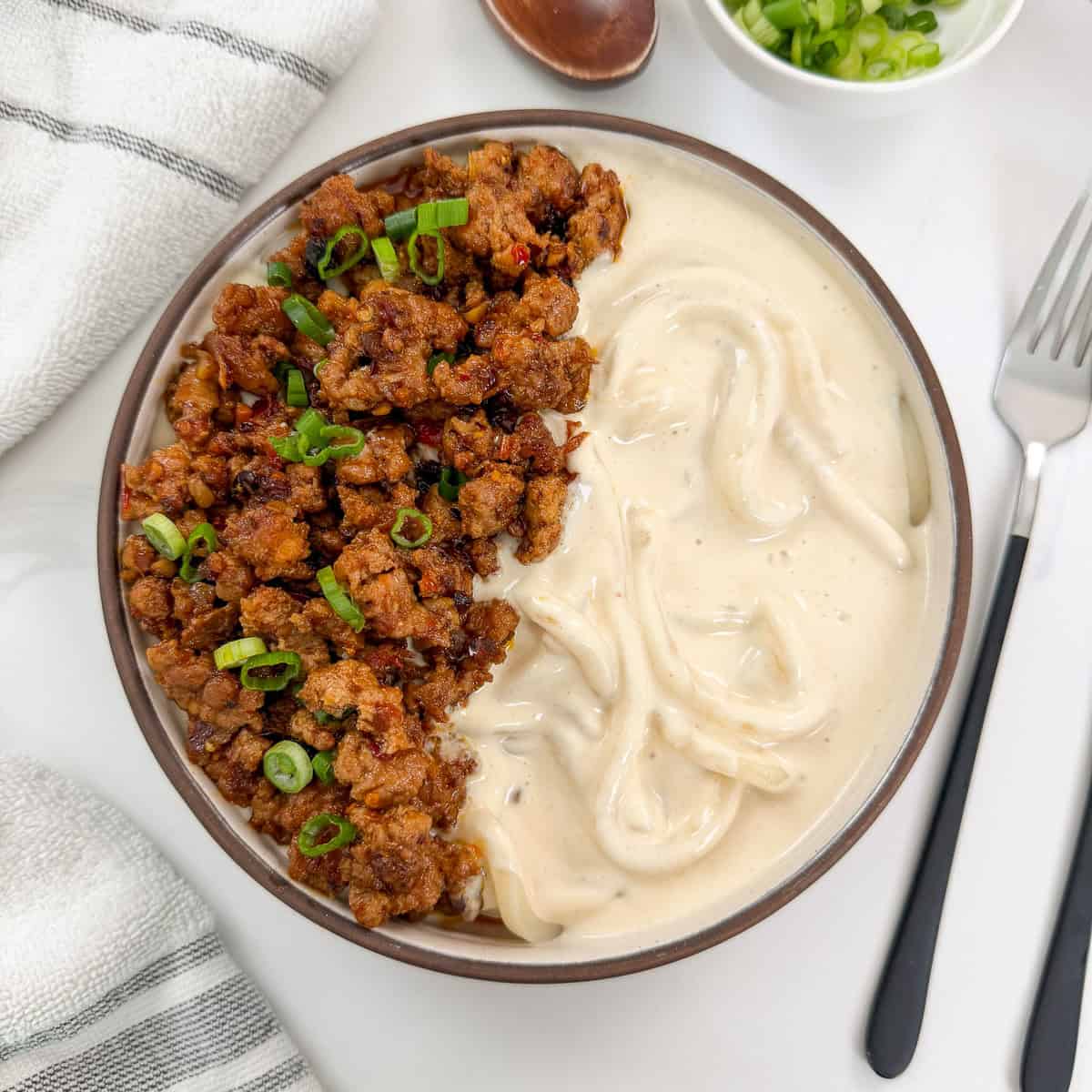 Creamy Mapo Tofu Udon in a bowl with minced meat and green onions on top. There is a fork on the right side and a wooden spoon above the bowl.