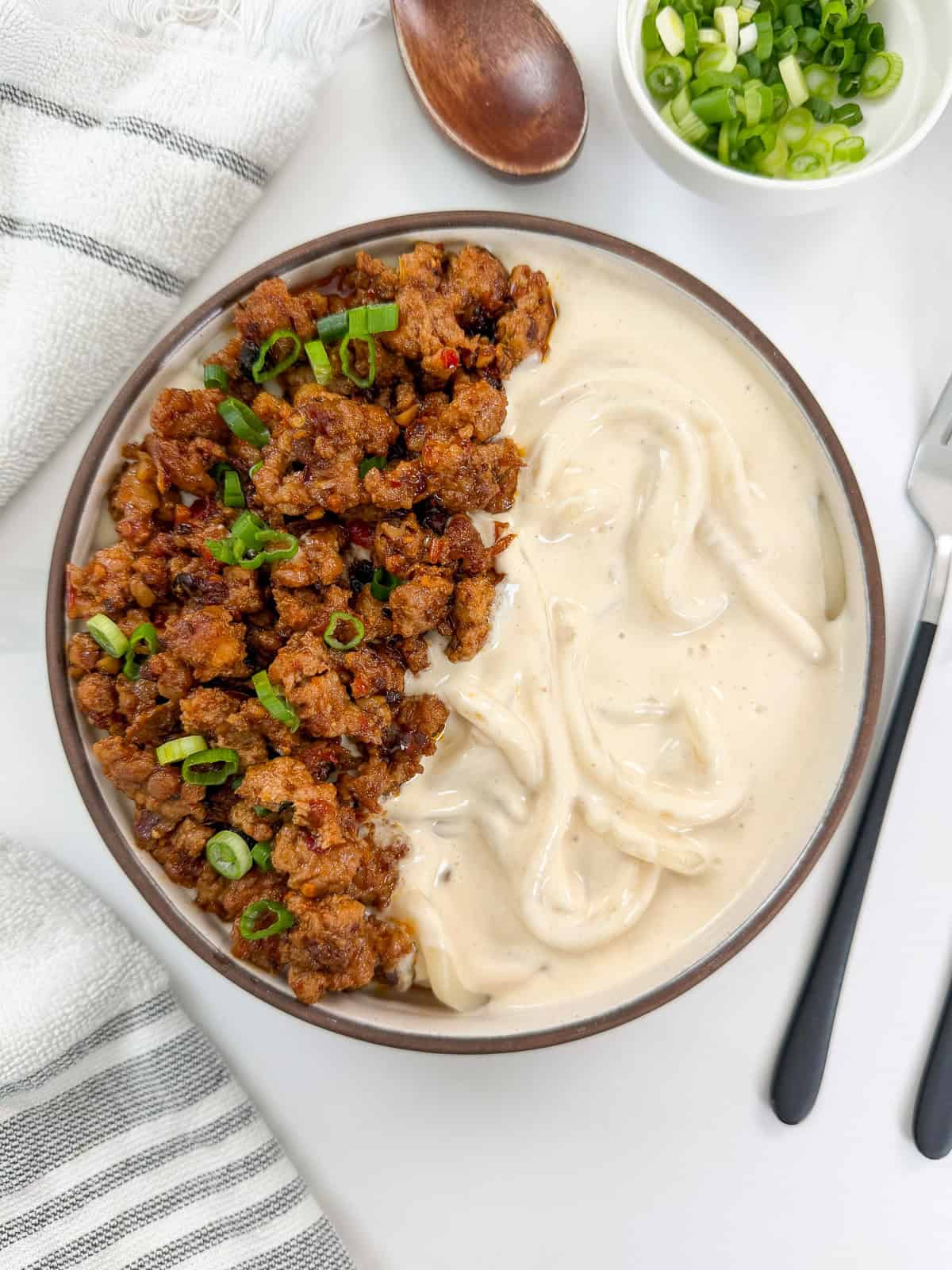Creamy Mapo Tofu Udon in a bowl with minced meat and green onions on top. There is a fork on the right side and a wooden spoon above the bowl and a towel to the left.