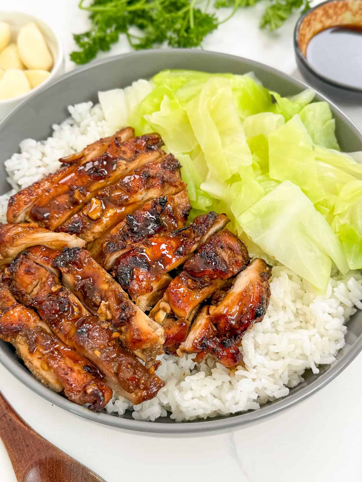 Five Spice Chicken in a bowl with white rice and cooked green cabbage. There is garlic, herbs and extra dipping sauce in the back.