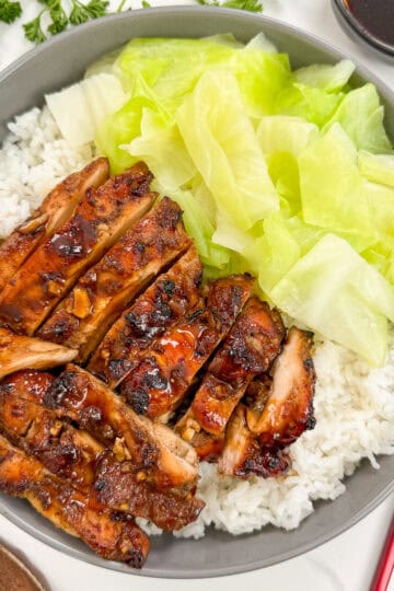 Five Spice Chicken in a bowl with white rice and cooked green cabbage. There is a brown spoon and red and gold chopsticks next to it. There is also garlic, herbs, and a side of dipping sauce on top.