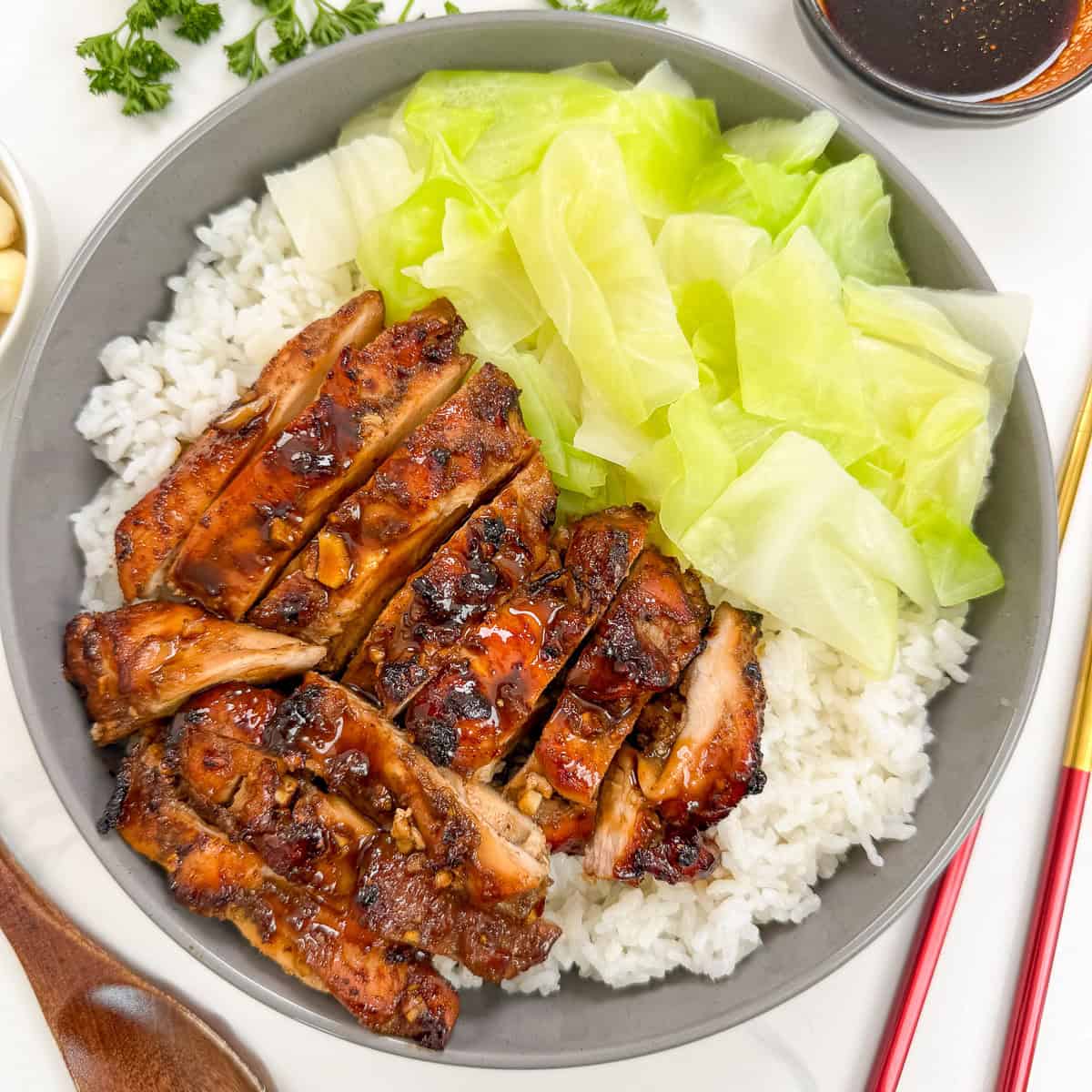 Five Spice Chicken in a bowl with white rice and cooked green cabbage. There is a brown spoon and red and gold chopsticks next to it. There is also garlic, herbs, and a side of dipping sauce on top.