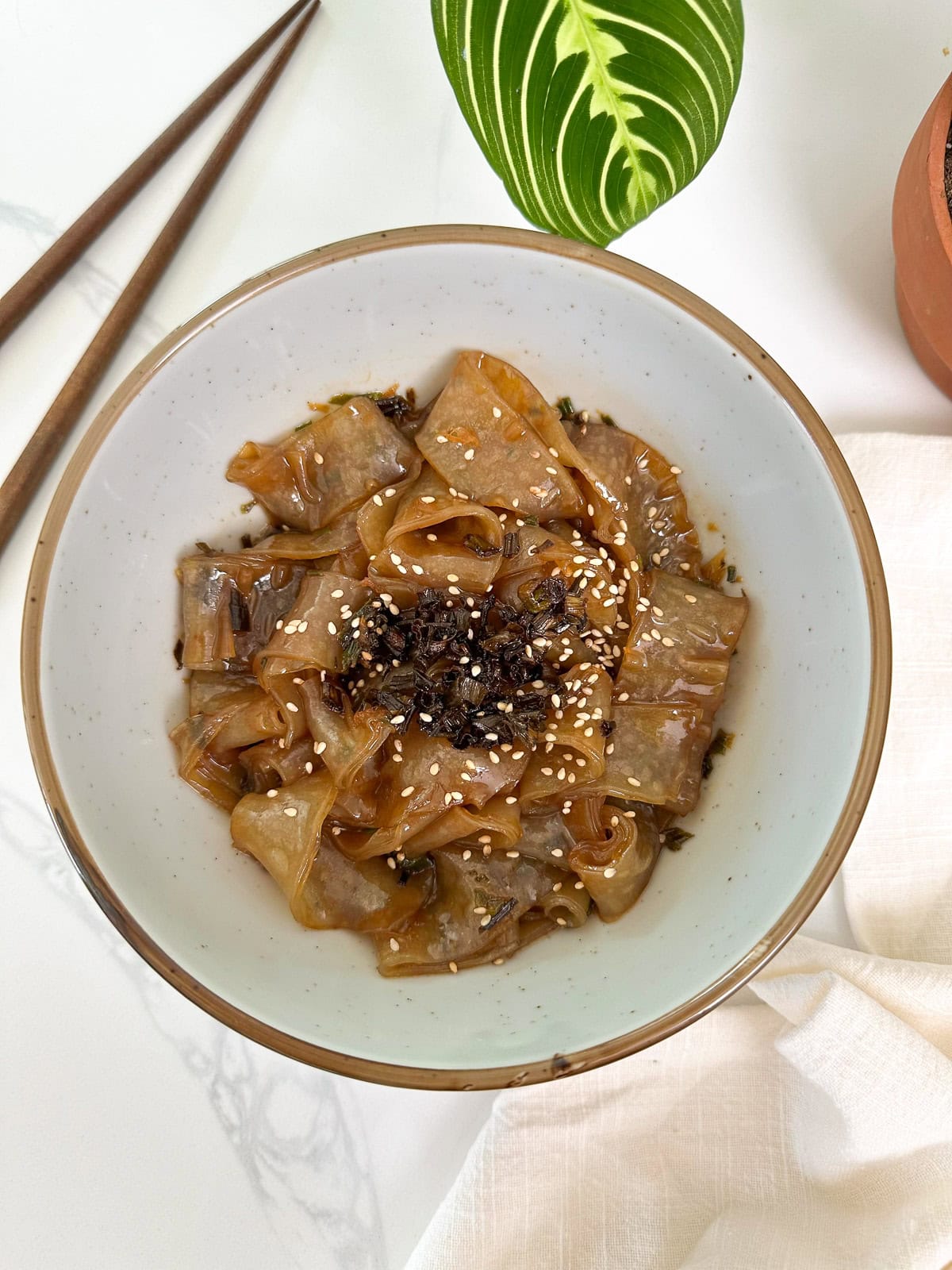 Scallion Oil Rice Paper Noodles in a bowl with chopsticks to the left and a plant at the top