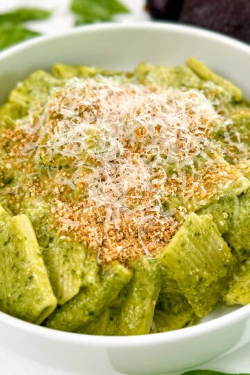 A white bowl filled with Avocado Pesto Pasta, coated in vibrant green sauce, topped with grated cheese and golden breadcrumbs, with fresh basil leaves in the background.