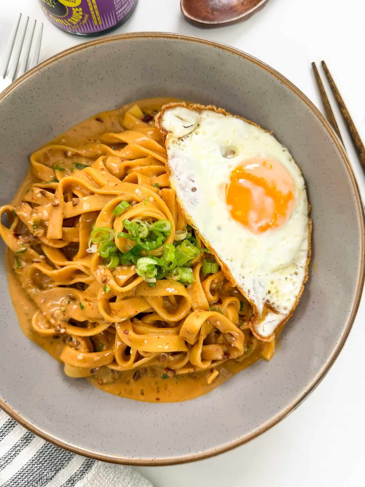 A bowl of Creamy Peanut Chili Noodles garnished with chopped green onions, served with a sunny-side-up egg on top. Chopsticks and a drink can are visible nearby.