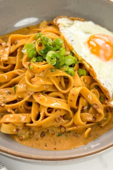 A bowl of creamy tagliatelle pasta reminiscent of Creamy Peanut Chili Noodles, topped with chopped green onions and served with a sunny-side-up fried egg on the side.