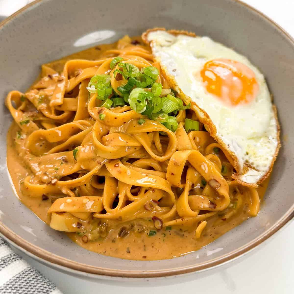 A bowl of creamy tagliatelle pasta reminiscent of Creamy Peanut Chili Noodles, topped with chopped green onions and served with a sunny-side-up fried egg on the side.