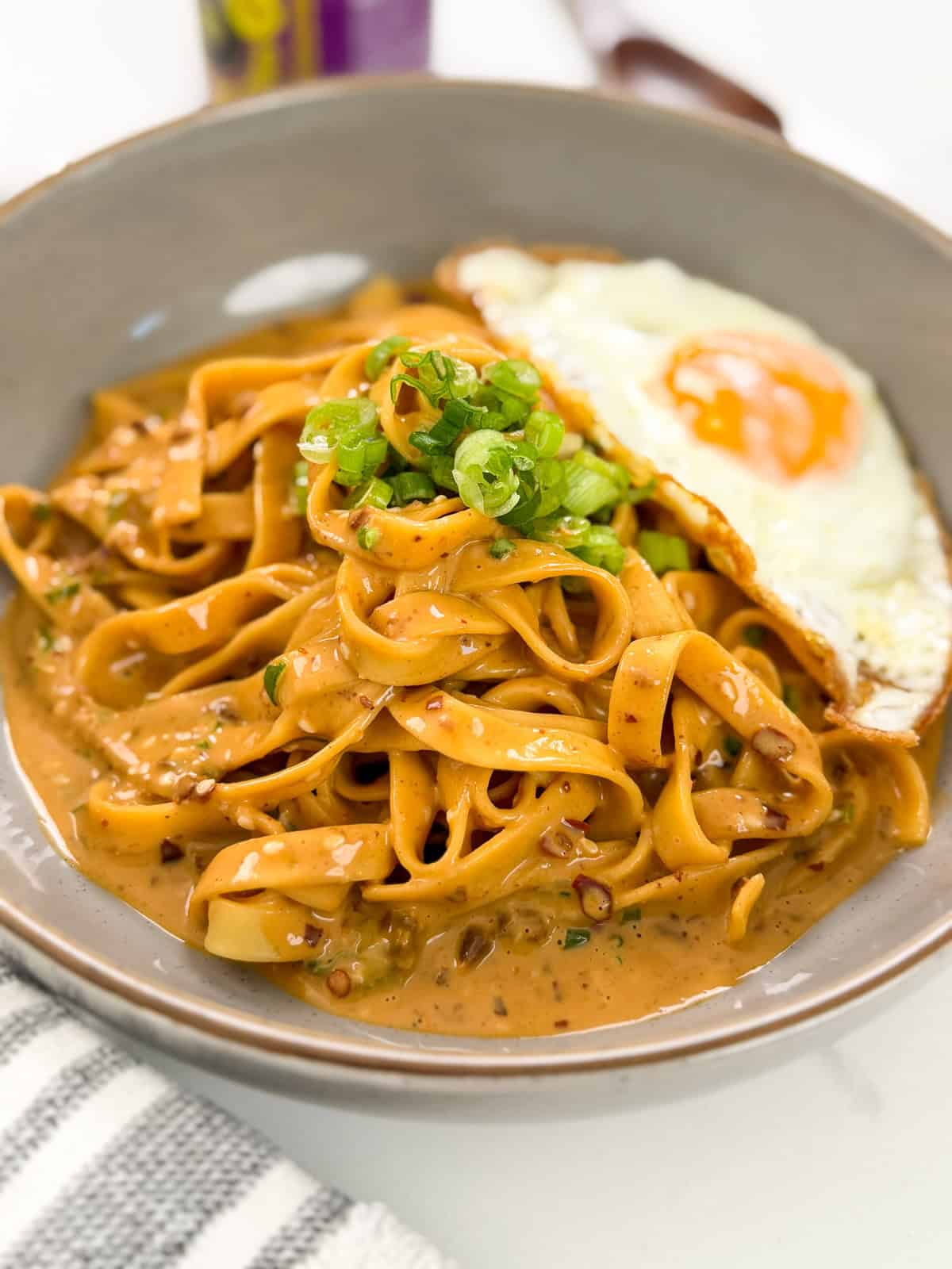 A bowl of Creamy Peanut Chili Noodles topped with chopped green onions and served with a sunny-side-up fried egg. A striped napkin is beside the bowl.
