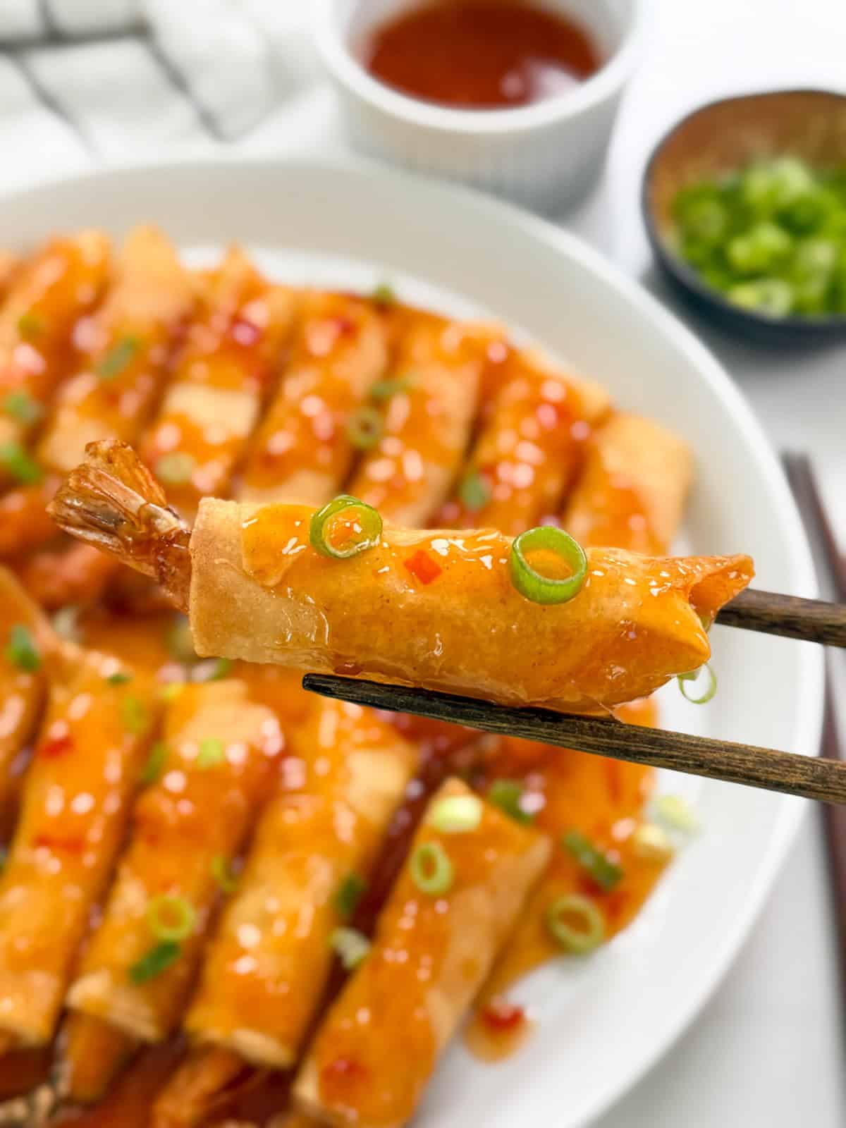 A close-up of chopsticks holding a crispy Firecracker Shrimp spring roll topped with sweet chili sauce and sliced green onions, with a plate of similar spring rolls in the background.