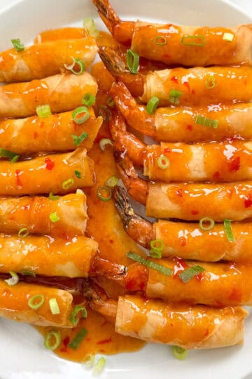 A plate of golden-brown Firecracker Shrimp spring rolls topped with a glossy sweet chili sauce and garnished with sliced green onions, arranged neatly on a white dish.