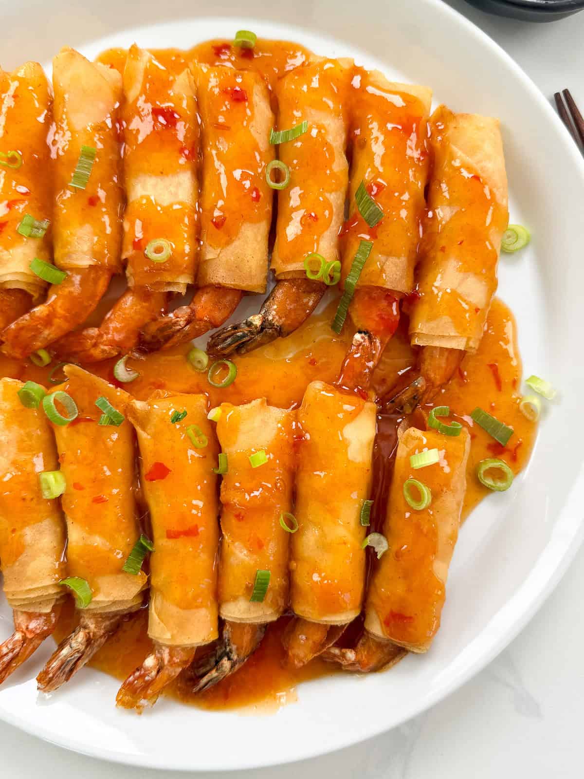 A plate of golden-brown Firecracker Shrimp spring rolls topped with sweet chili sauce and garnished with chopped green onions.