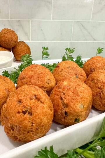 Golden-brown hush puppies and crispy Fried Tuna Balls arranged on a white tray, garnished with fresh parsley, with two small bowls of dipping sauce and more bites in the background. White tiled wall behind the food.