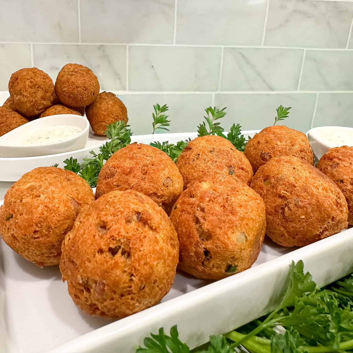 Golden-brown hush puppies and crispy Fried Tuna Balls arranged on a white tray, garnished with fresh parsley, with two small bowls of dipping sauce and more bites in the background. White tiled wall behind the food.