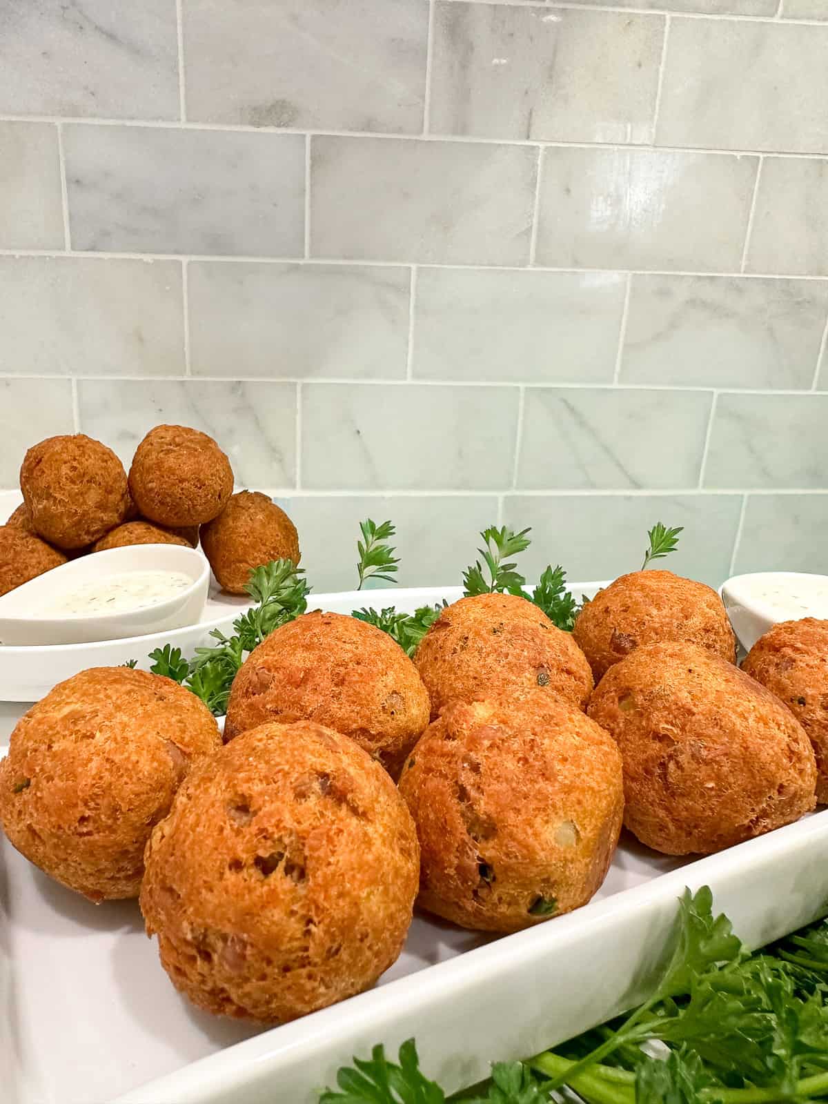 A tray of golden-brown, round Fried Tuna Balls garnished with fresh parsley, with small bowls of dipping sauce and extra hush puppies in the background, set against a tiled kitchen wall.