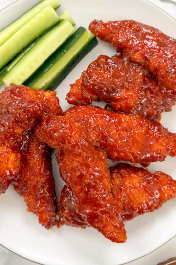A white plate with several pieces of glazed Honey BBQ Chicken Tenders and a side of cucumber sticks.
