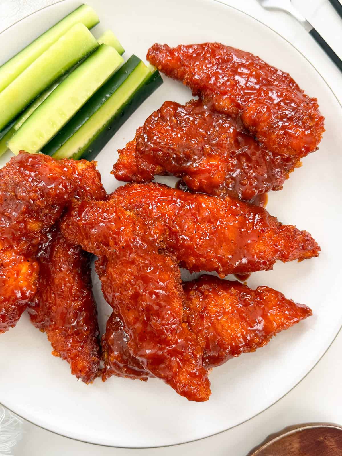 Plate with crispy, saucy Honey BBQ Chicken Tenders coated in a reddish glaze, served alongside fresh cucumber sticks.