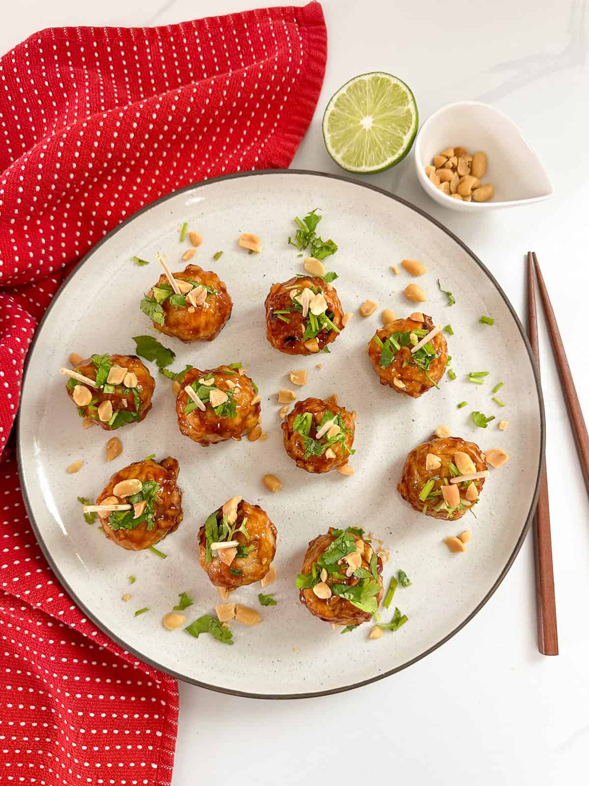 A plate of ten Kung Pao Chicken Meatballs garnished with chopped peanuts and cilantro, each with a toothpick, sits next to chopsticks, a halved lime, a small bowl of peanuts, and a red dotted cloth napkin.