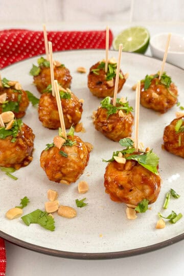 A white plate with nine Kung Pao Chicken Meatballs on toothpicks, glazed and garnished with chopped peanuts and cilantro. A red napkin, lime half, and small white bowl are in the background on a white surface.