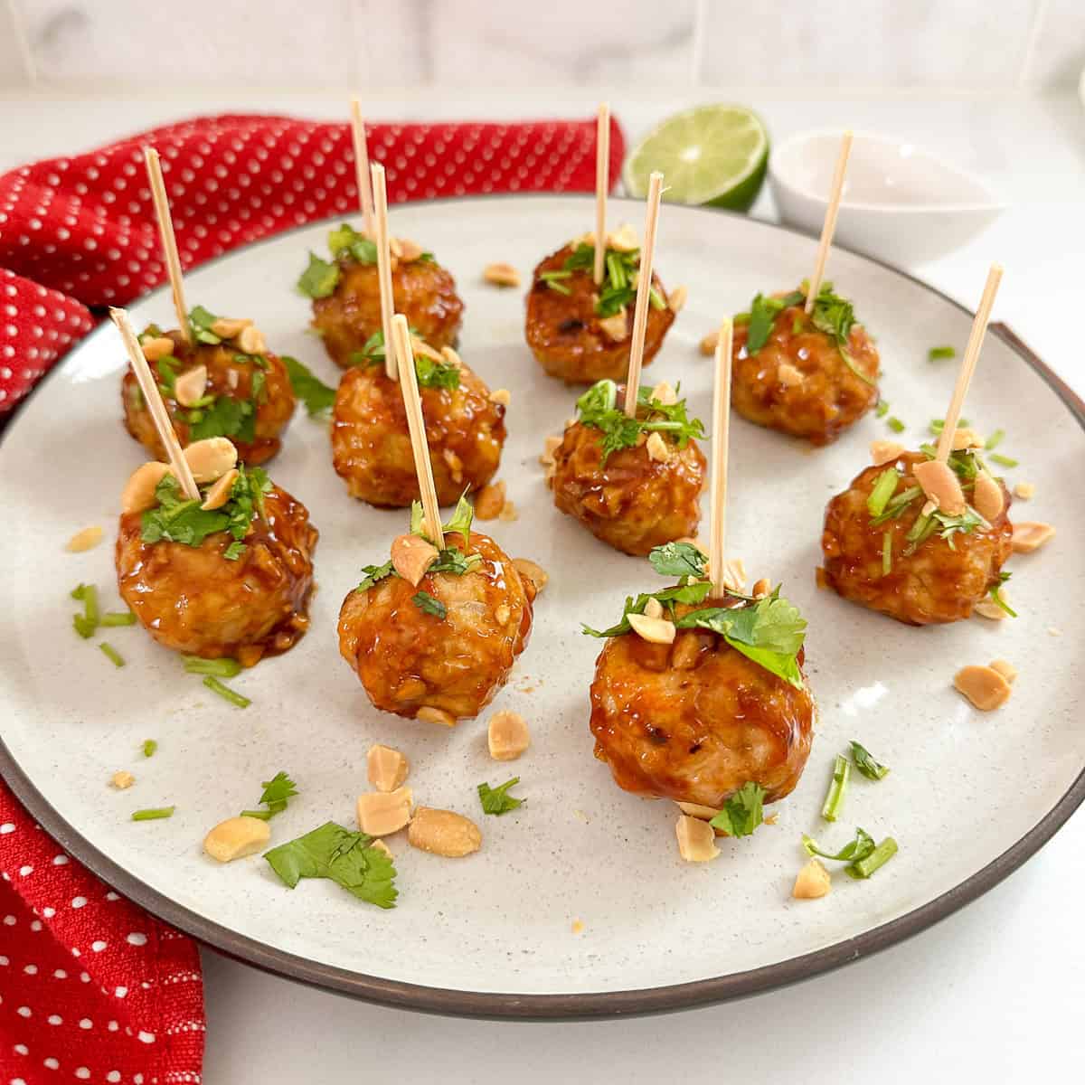 A white plate with nine Kung Pao Chicken Meatballs on toothpicks, glazed and garnished with chopped peanuts and cilantro. A red napkin, lime half, and small white bowl are in the background on a white surface.