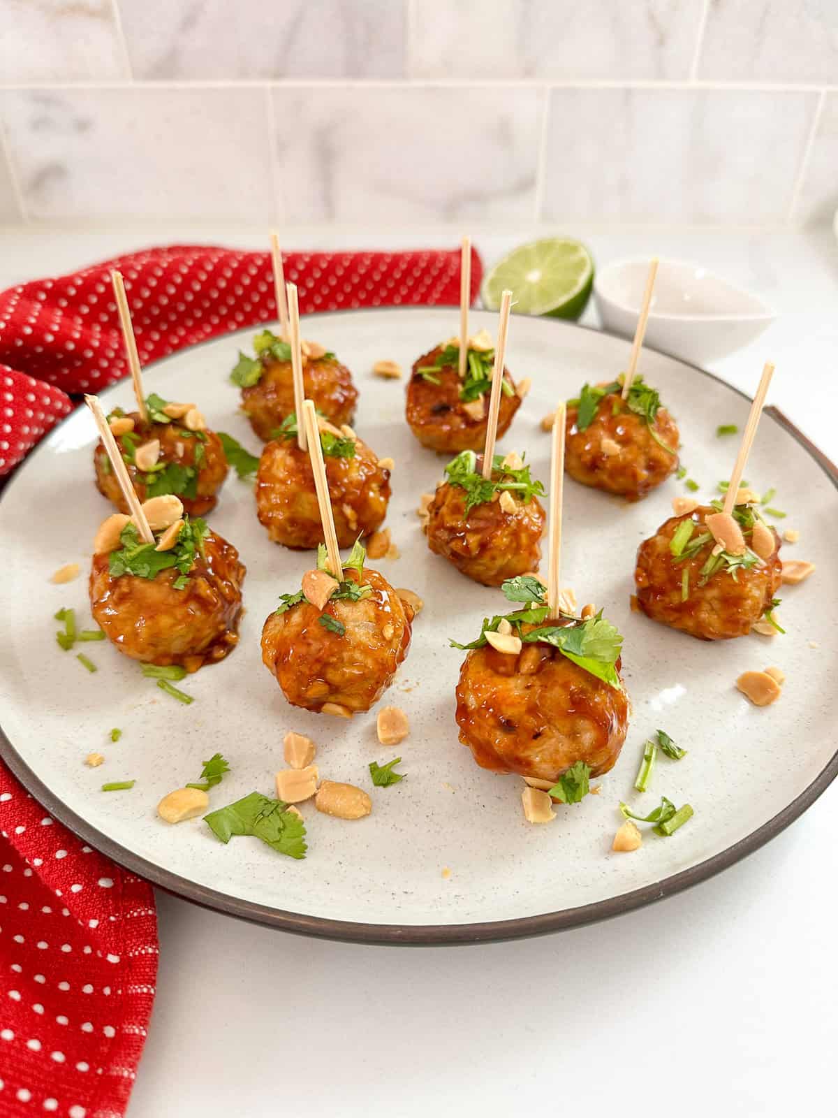 A plate of Kung Pao Chicken Meatballs garnished with chopped peanuts and cilantro, each pierced with a toothpick. A red cloth, lime half, and small white bowls are in the background on a white countertop.