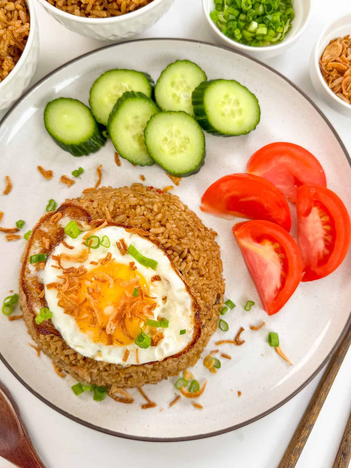 Nasi Goreng (Indonesian Fried Rice) on a plate with fried egg on top, fried shallots and a side of tomato wedges and sliced cucumbers.