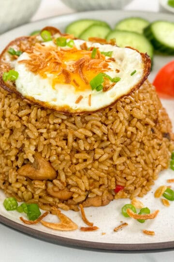 Nasi Goreng (Indonesian Fried Rice) on a plate with green onions, crispy fried shallots, and a fried egg on top. There are tomato wedges and sliced cucumbers on the side.
