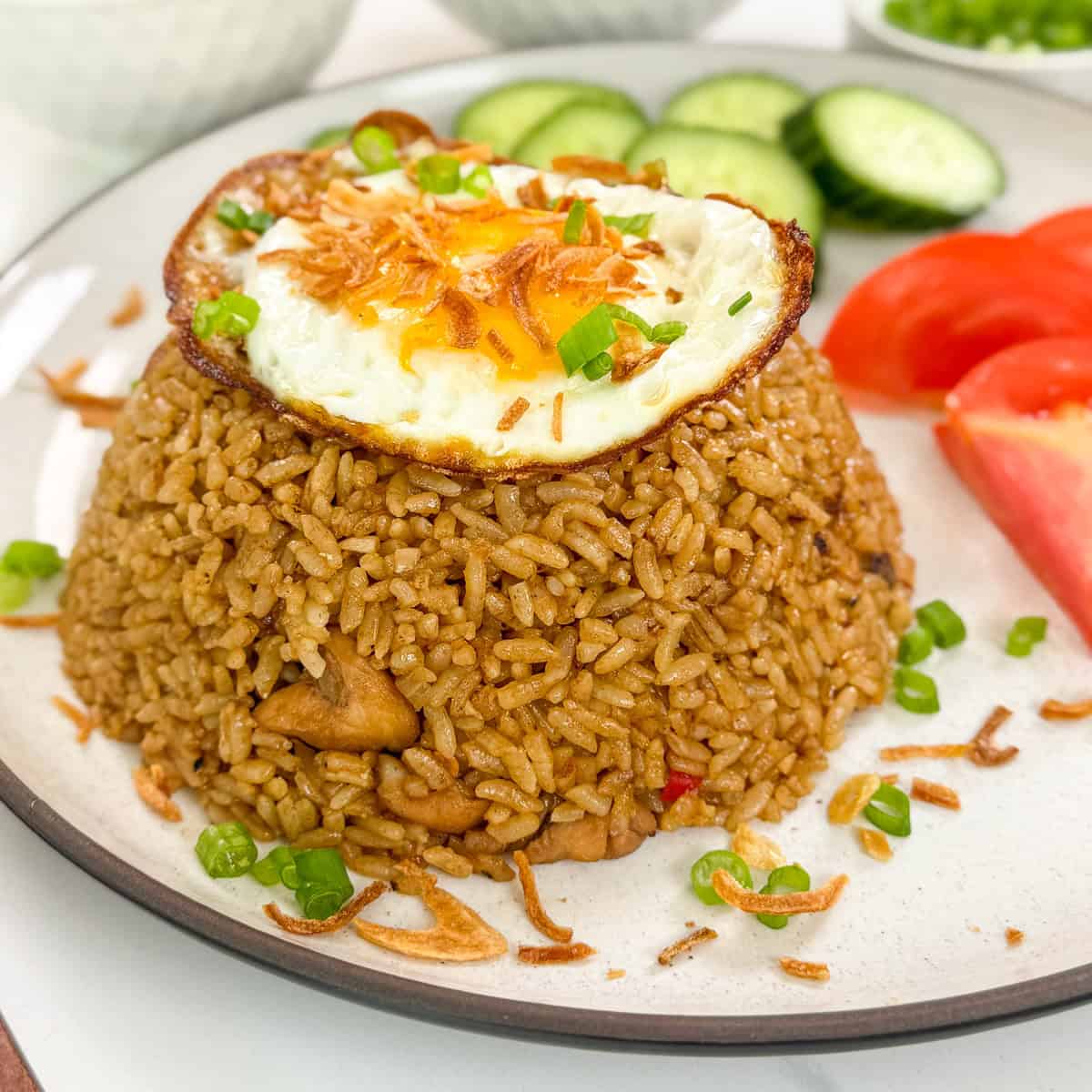 Nasi Goreng (Indonesian Fried Rice) on a plate with green onions, crispy fried shallots, and a fried egg on top. There are tomato wedges and sliced cucumbers on the side.