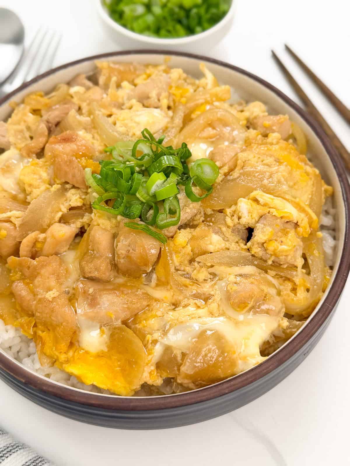 A bowl of Oyakodon (Japanese Chicken and Egg Rice Bowl) features tender chicken, onions, and scrambled eggs over rice, garnished with sliced green onions. Chopsticks and a small bowl of extra green onions are visible in the background.