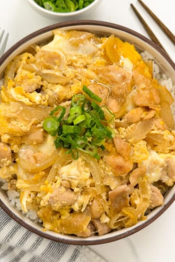 A bowl of Oyakodon (Japanese Chicken and Egg Rice Bowl), topped with tender chicken, onions, and scrambled eggs, is garnished with chopped green onions. Chopsticks, a spoon, and a small side bowl of green onions accompany the dish.