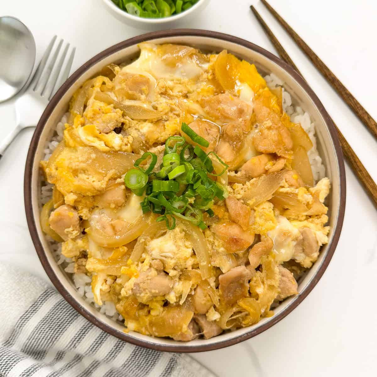 A bowl of Oyakodon (Japanese Chicken and Egg Rice Bowl), topped with tender chicken, onions, and scrambled eggs, is garnished with chopped green onions. Chopsticks, a spoon, and a small side bowl of green onions accompany the dish.
