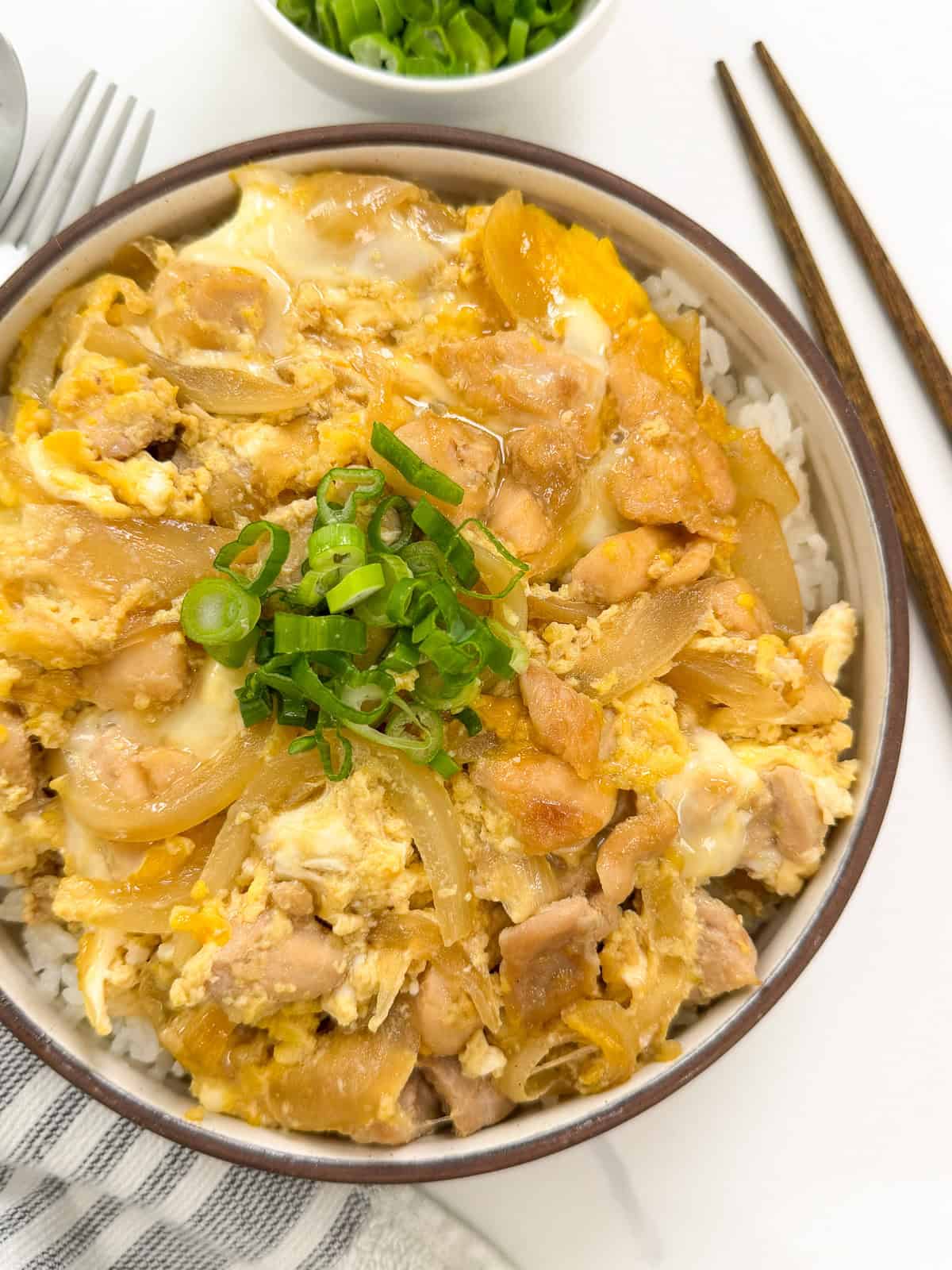 A bowl of Oyakodon (Japanese Chicken and Egg Rice Bowl) featuring tender chicken, onions, and scrambled eggs over rice, topped with sliced green onions. Chopsticks rest beside the bowl on a white surface.