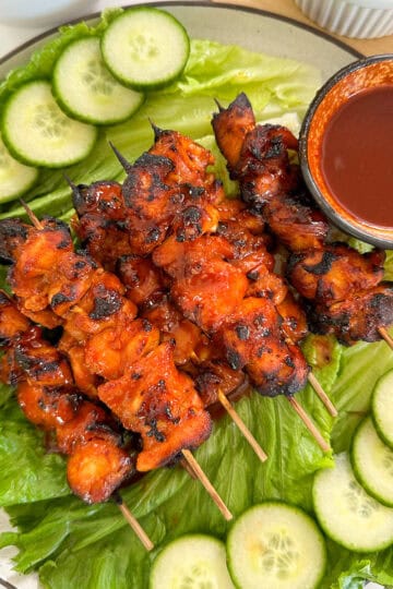 Spicy Gochujang Chicken Skewers are coated in a bold red sauce, arranged on a bed of lettuce, with sliced cucumbers on the side and a small bowl of dipping sauce served alongside.