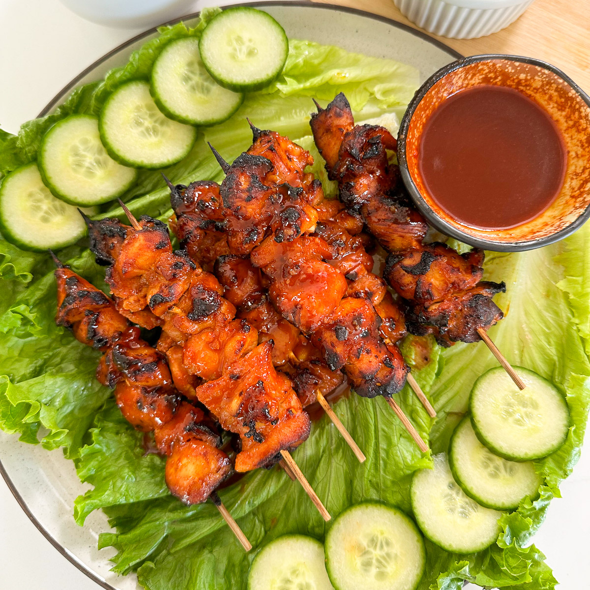 Spicy Gochujang Chicken Skewers are coated in a bold red sauce, arranged on a bed of lettuce, with sliced cucumbers on the side and a small bowl of dipping sauce served alongside.